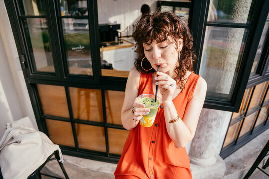 Relaxed middle-aged woman drinking iced matcha outside specialty cafe, high angle view