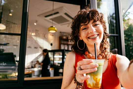 Happy middle-aged woman with braces taking selfie with iced matcha outside modern cafe