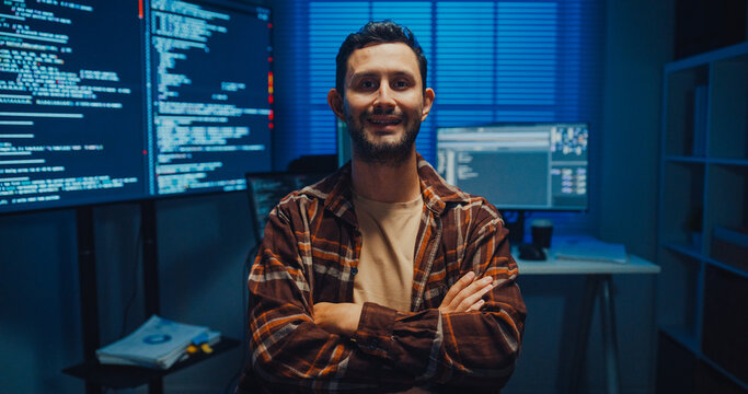 Portrait of confident Caucasian male programmer smiles looking at camera in office at night. Professional developer workspace, Secure computer network system, Advanced cybersecurity data concept.