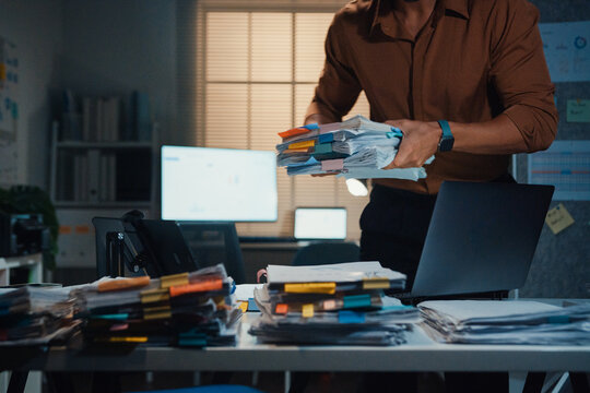 Stressed young Latin businessman walking to place tax document stack at desk in dark night office. Overworked overtime, heavy workload stress, tired employee, corporate business burnout concept.