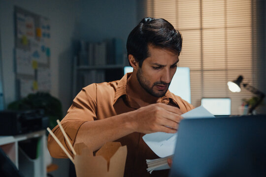 Young Latin businessman eating takeout instant noodles and reviewing document on laptop at desk in dark night office. Overtime working late, overworked deadline, fast food, corporate problem concept.