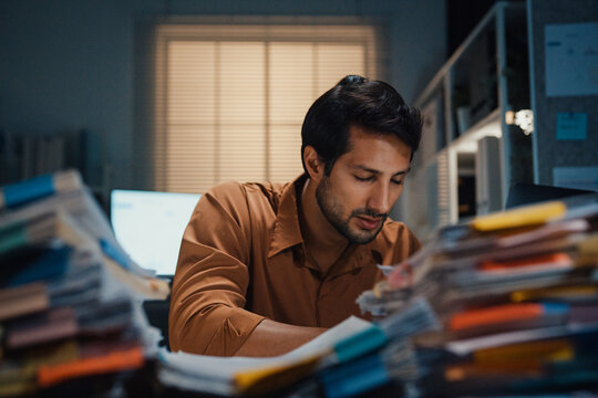Sleepy young Latin businessman yawning over heavy paperwork stack at desk in dark night office. Overwork burnout, working late overtime, corporate job stress, exhausted professional manager concept.