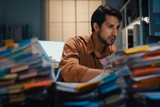 Overworked young Latin businessman working late on document in large stack of paperwork files at office night. Tax audit deadline, accounting stress, corporate burnout, urgent business concept.