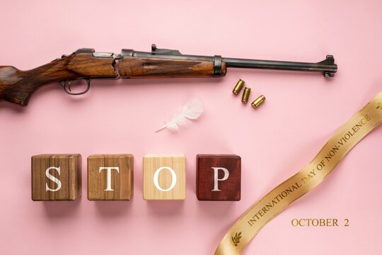International Day of Non Violence Concept Flat Lay with Hunting Rifle, Bullets, Feather, Wooden Blocks Spelling STOP, and Ribbon on Pink Background