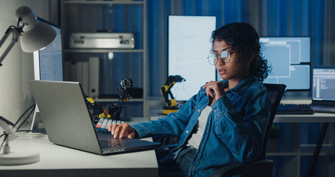 Young African American female programmer coding AI software on multiple screens in night office. Network security, cybersecurity, machine learning, working late, overtime, robotics automation concept.