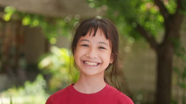 Portrait of adorable Asian kid looking directly at the camera with a gentle smiling face in a red shirt standing outdoors with beautiful green bokeh background. Childhood happiness lifestyle concept.