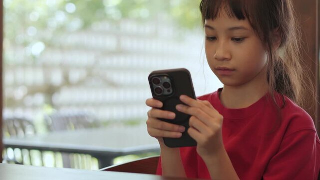 Asian kid using a smartphone at restaurant. Cute Thai girl excitedly watching video or playing games. Shot with selective focus and beautiful copy space for text. Childhood digital lifestyle concept.
