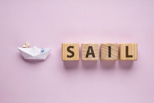 Word SAIL spelled with wooden cube blocks next to paper boat on pink background business startup concept