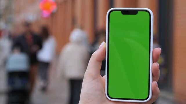 Hand holds smartphone with green screen. Background shows blurred pedestrians on street. Suitable for app mockups or digital content. Urban setting with natural daylight. Ideal for advertising