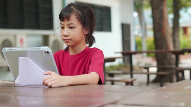 Asian kid using stylus pen on a digital tablet for e-learning at outdoor school table. Selective focus shows the young student engaging with technology for modern education and digital study concepts.