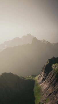 At dawn, the sun rises over the mountains, casting light on the peaks and illuminating the fog. Green hills are visible in the foreground, creating a natural landscape.