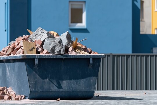 Large metal construction skip bin filled with bricks and concrete debris in front of a blue house for demolition services and renovation waste hire