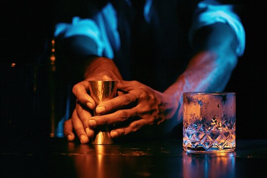 Close up of a mans hands holding a brass jigger beside a crystal rocks glass on a dark bar for premium cocktail branding and hospitality concepts