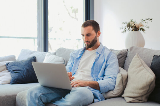 Young man sits on a couch using a laptop at home, focused and relaxed while working remotely in a bright, modern living room with soft cushions and natural light.