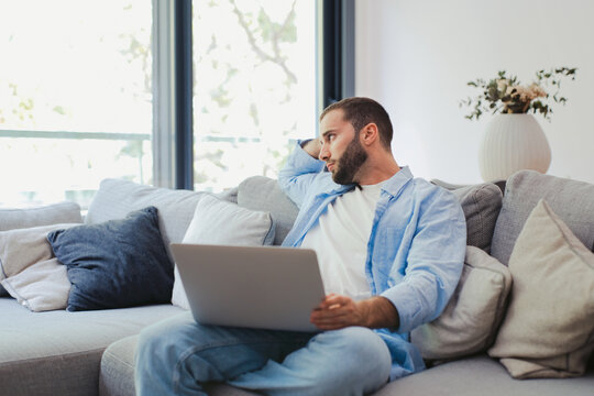 Young man with a beard and a casual shirt is sitting on a comfortable sofa with a laptop on his lap, looking away and reflecting, possibly contemplating remote work or experiencing procrastination