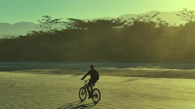 Silhouette of cyclist riding on a reflective beach during golden sunrise. Peaceful coastal scene with warm sunlight and active lifestyle. 4K video.