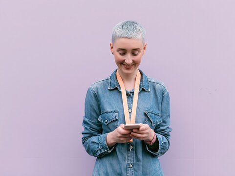Smiling woman with short cropped hair using a smartphone against a lilac background, wearing a denim shirt and pink lanyard for mobile app and lifestyle technology imagery