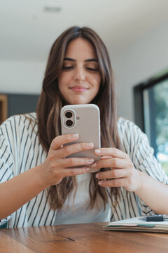 Young woman sits at a home desk, smiling as she uses her smartphone to text, browse social media, and manage remote work tasks. Casual lifestyle scene with natural light and a notebook nearby.