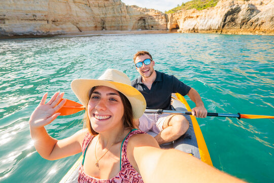 Young couple enjoying a summer adventure, paddling an inflatable kayak in turquoise ocean water along a scenic rocky coast and taking a smiling selfie