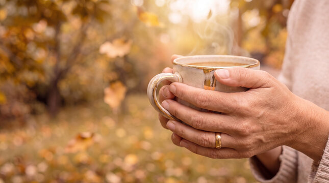Woman hands holding warm mug in a cozy sweater against a blurred autumn forest. Close up lifestyle concept of seasonal comfort and relaxation