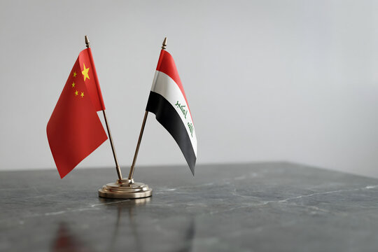 Miniature flags of china and iraq displayed on a dark marble table against a plain light gray wall for international cooperation concepts