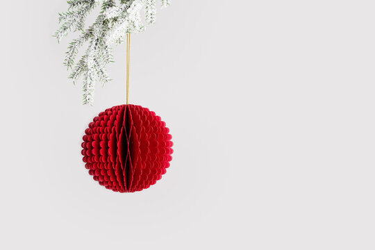 Christmas paper bauble hanging on golden string, close up red round ball on snow white fir tree branch on grey background, copy space. New Year Winter holiday minimal design greeting card, invitation