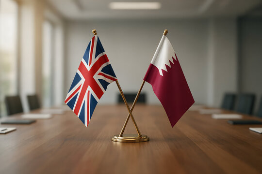 Two small desk flags of the united kingdom and qatar standing together on a wooden conference table inside a blurred corporate meeting room