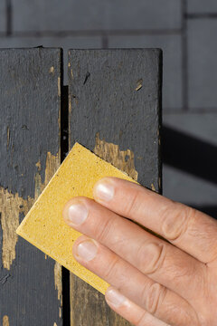 Man sanding loose peeling black paint off a wooden outdoor patio table. Garden DIY chore concept.