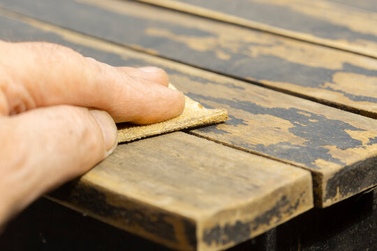 Man sanding loose peeling black paint off a wooden outdoor patio table. Garden DIY chore concept