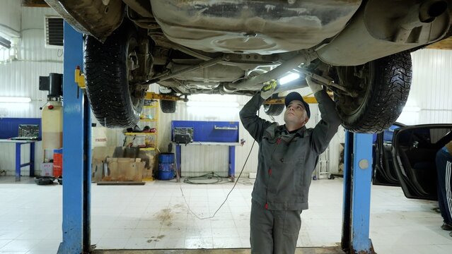 Experienced mechanic checking vehicle undercarriage with inspection light, working on hydraulic ramp in modern auto repair workshop, performing detailed diagnostic and maintenance procedures