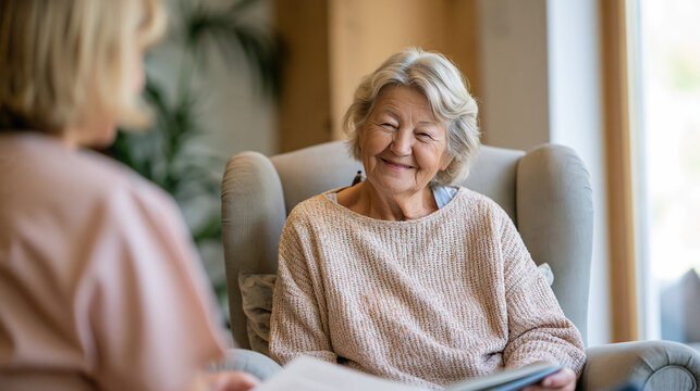 An elderly person sitting comfortably in a well-lit living room armchair, while a caregiver sits nearby, reading a book aloud or sharing a conversation.