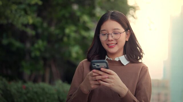 Happy Asian businesswoman wearing eyeglasses using smartphone outdoors near office park during sunset, cheerful female entrepreneur smiling while texting on mobile phone online app for hybrid work dig