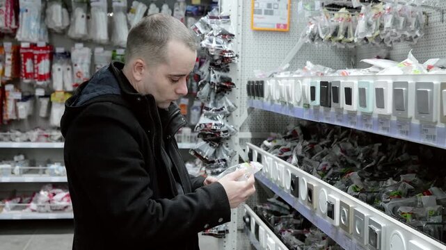 Skilled electrician selecting light switches, comparing various models in hardware store aisle, preparing for upcoming electrical installation project