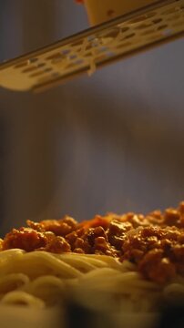 A chef grates cheese over a plate of spaghetti bolognese using a grater, preparing a meal with the dish and glassware set on a table, in a home kitchen, creating an inviting atmosphere