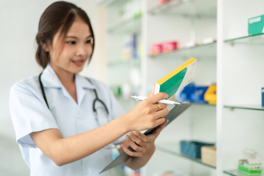 A woman in a white lab coat is holding a clipboard and looking at a box of pills