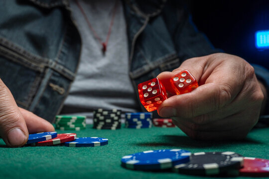 A person holds two red dice in one hand while poker chips are arranged on a green table. The scene shows an indoor setting focused on a game of chance during the evening
