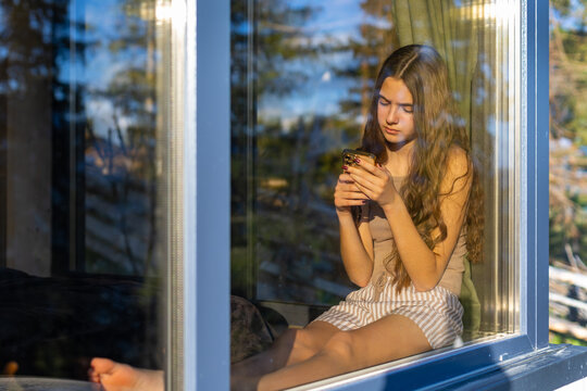 Young Woman Using Smartphone Beside a Sunlit Window. Young woman sitting beside a window and using a smartphone in warm afternoon sunlight.