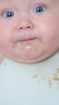 Closeup of a cute baby with big blue eyes getting a messy face while a parent feeds them their first solid food, a fruit puree from a spoon, an important milestone in childhood development