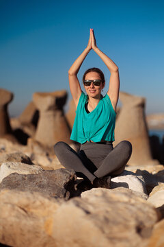 Healthy Woman Doing Yoga Meditation Sitting In Lotus Pose On Wave breaker. Peaceful sporty girl practices mindfulness exercises while keeping arms raised near sea shore