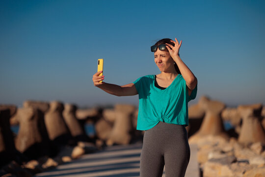 Young Woman Lifting Sunglasses Squinting Her Eyes Taking Funny Selfie. Cheerful female person raises eyewear while looking at phone camera making self-portrait