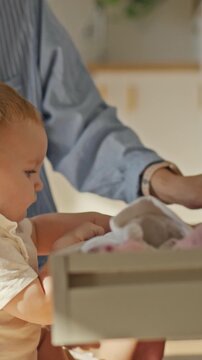 A mother and child fold laundry together in a bright kitchen, sorting shirts and pants into closets while a housewife arranges other items, a routine of household organization and parenting