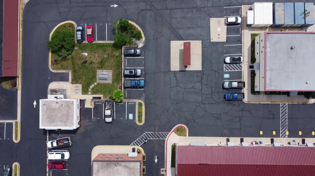 Top-down drone shot of a commercial parking lot and drive-thru. Features parked cars, cracked asphalt, painted arrows, and small retail buildings in a suburban shopping center in Fairfax, Virginia.