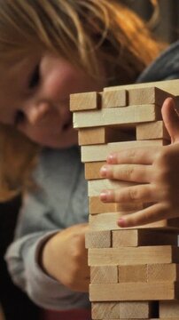 A mother and her young daughter sit on the home floor, playing a jenka game as they stack wooden blocks into a tower, a family activity captured in a relaxed evening setting with soft lighting