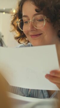 A young designer in a well-lit studio sits at a cluttered table, reviewing sketches and handwritten notes while fabric samples and sewing tools lie nearby, indicating a design development process