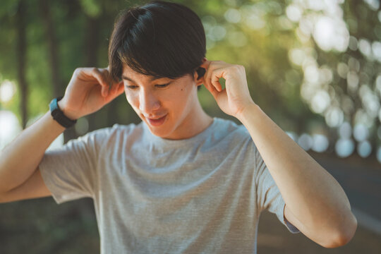 Close up of asian male putting in ear wireless earphones getting ready for workout standing on public park in summer morning.