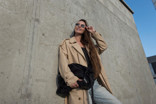 Urban trendy shot of a smiling young woman with long hair wearing a beige trench coat and sunglasses, holding a black bag against a concrete wall under a blue sky. Urban street fashion.
