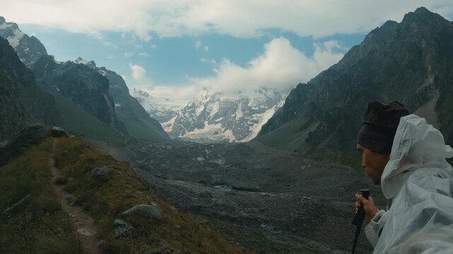 A male hiker wearing a white raincoat and hat walks along a grassy trail along a ridge, overlooking a vast rocky moraine valley and distant snow-capped glaciated peaks under a cloudy sky
