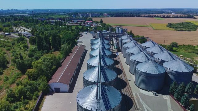 Dynamic aerial overview reveals long rows of silver metal silos at modern agricultural grain elevator facility near countryside fields under blue sky.