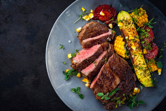 Dry aged roasted beef entrecote with grilled vegetables, zucchini, sweet corn and greens served as top view on a design plate with copy space right