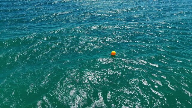 A bright orange buoy floats on the surface of the sea. Waves move around it as sunlight shines down. The scene shows clear blue water and bright conditions.